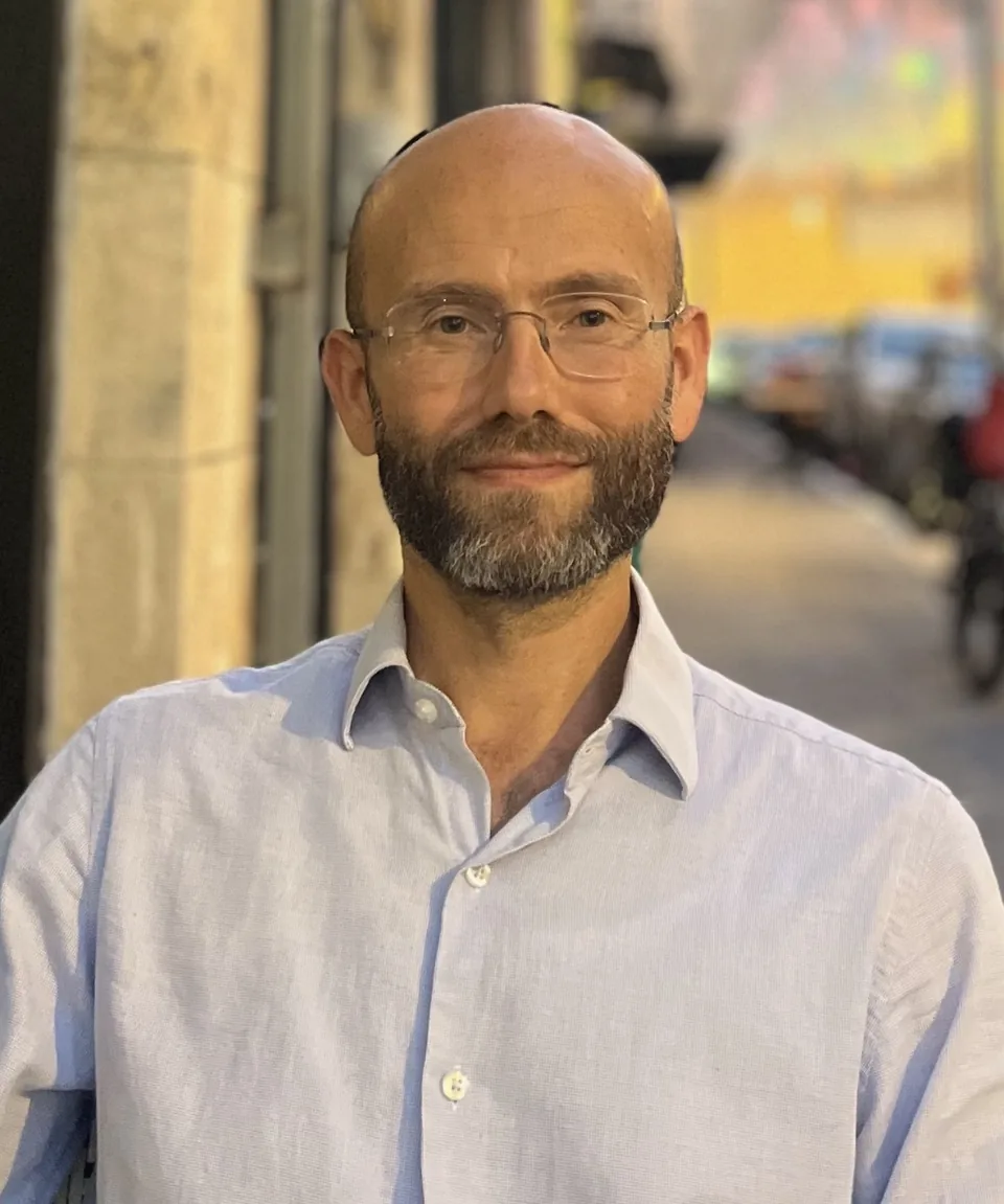 Betzalel Lapidus, bearded and wearing glasses and a pale blue linen shirt, looking to camera on a sunlit Jerusalem street.